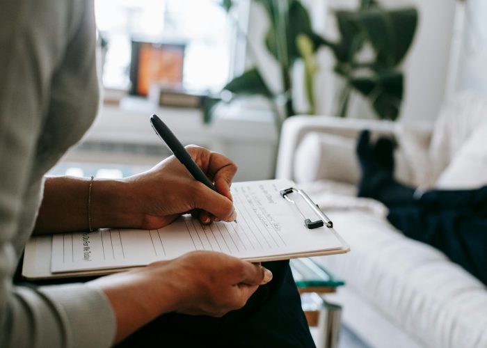 Unrecognizable ethnic female therapist taking notes on clipboard while filling out form during psychological appointment with anonymous client lying on blurred background